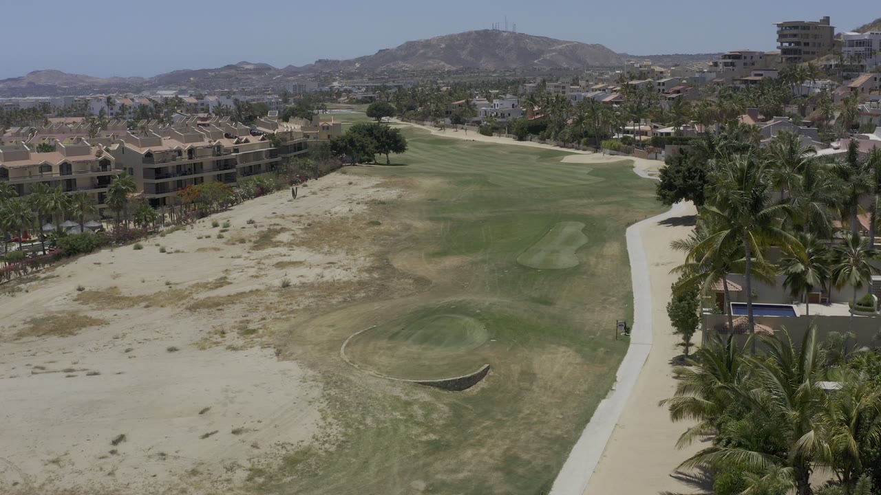 Aerial view in Los Cabos, Mexico, with a broad green fairway-like area between sandy patches, palm trees, buildings, and distant hills, with no visible people.