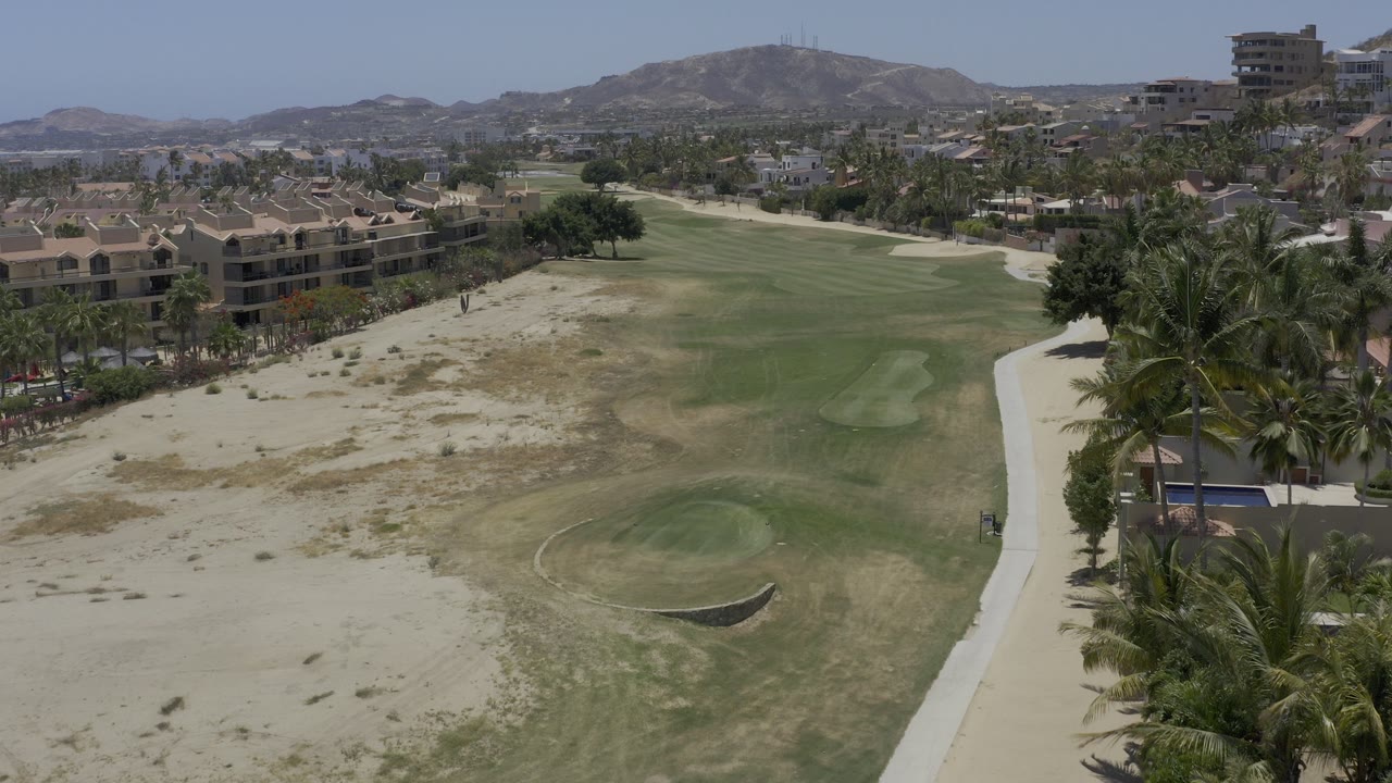 Aerial view over a sandy Los Cabos golf course with a green fairway, a rounded foreground green, resort buildings at left, palms at right, distant hills, and no visible people.