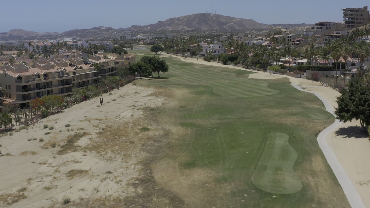 Aerial frame over a Los Cabos golf fairway with sandy rough in the foreground, low residential buildings at left, distant hills, and no visible people.