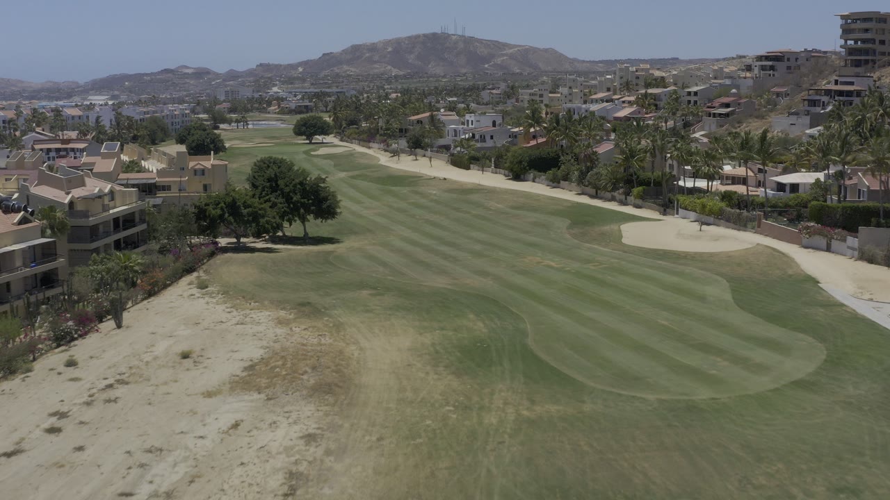 Aerial view of striped golf fairway, sand trap, palms, buildings, and hills.