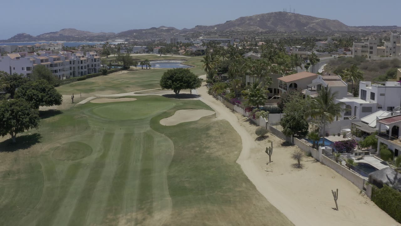 Aerial Los Cabos frame with striped fairway grass, curved sand bunkers, resort-style buildings, palms, dry hills, and no visible people.