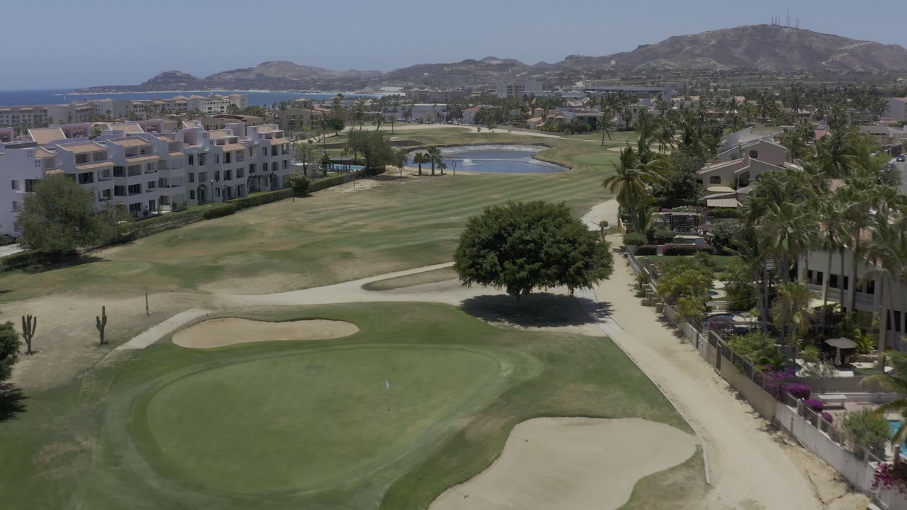 Elevated view of a golf green, sand bunkers, villas, palms, hills, and distant water.