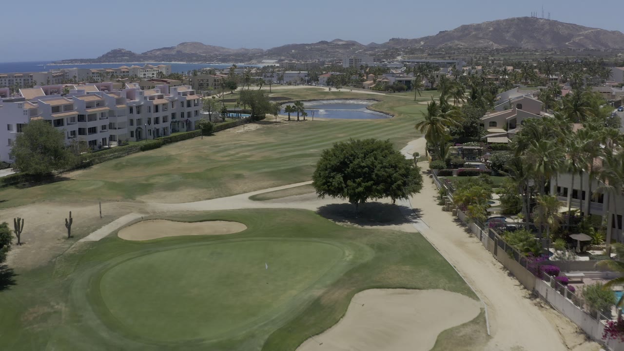 Los Cabos aerial view holds over manicured fairways, a foreground green, sand traps, white villas, palms, distant hills, blue water, and no visible people.