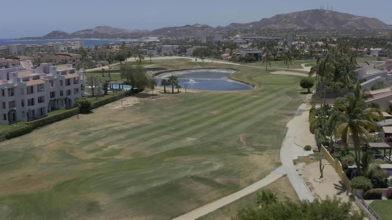 Elevated view over a grassy fairway, small lake, palms, buildings, water, and hills.