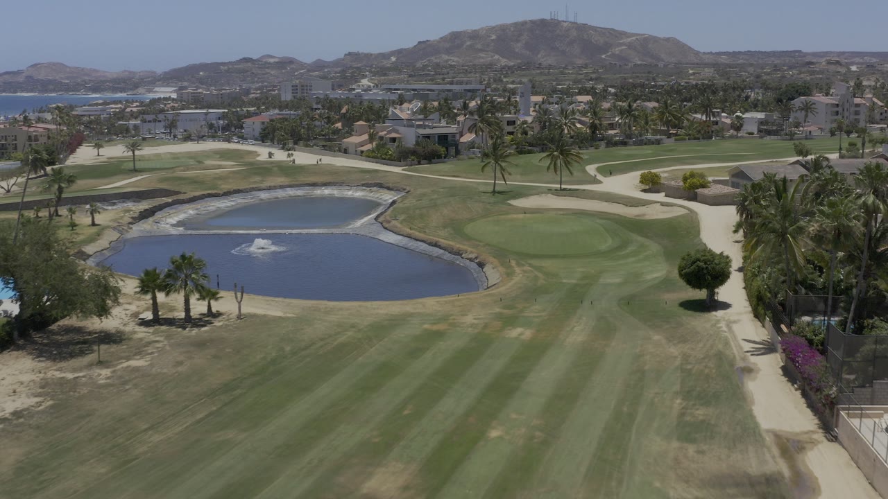 Elevated view of a country club lake, fountain, palms, lawns, and distant hills.