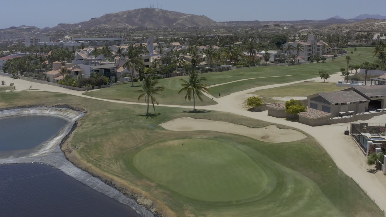 Aerial view of a golf green beside water, sand bunkers, palms, homes, and distant hills.