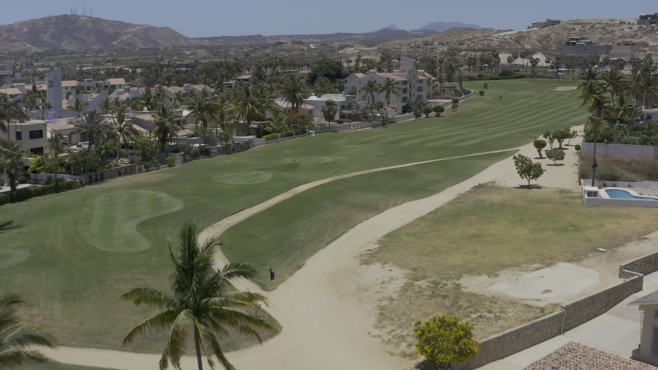 Aerial view of a green fairway, sand path, palms, houses, and dry hills.