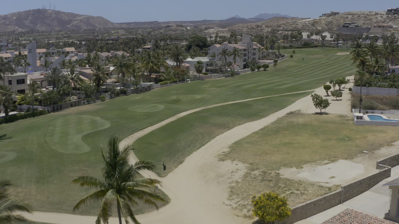 Second aerial frame in Los Cabos, Mexico, with the curving sand path more centered between manicured green grass, palms, residential buildings, and distant hills with no clearly visible people.
