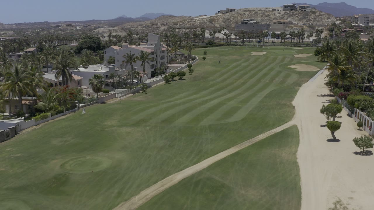 Aerial frame from Los Cabos, Mexico showing a striped golf fairway, pale sand path, modern homes, palms, desert hills, and tiny distant people on the course.