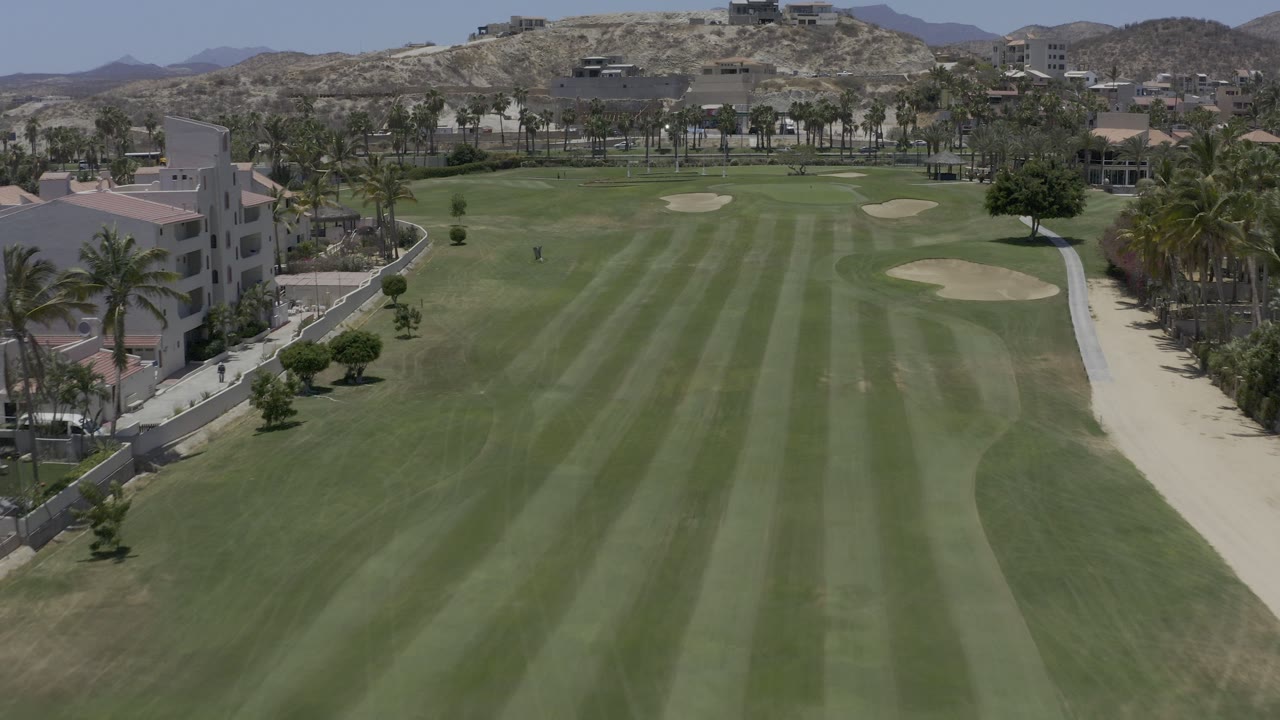 Aerial view down a green golf fairway with homes, palms, bunkers, and dry hills.