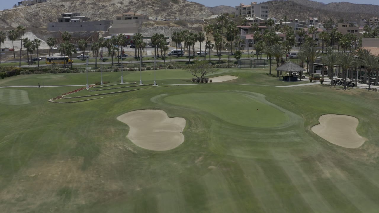 Wide elevated view in Los Cabos, Mexico, looking over manicured green turf, two sand traps, palm-lined road traffic, hillside buildings, and no visible people.