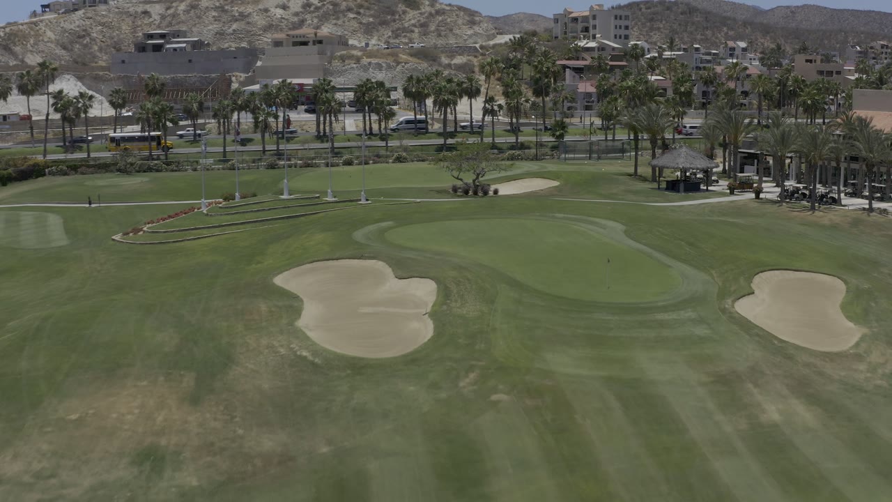 Elevated Los Cabos view with the central green and sand traps framed by white flagpoles, palm trees, a yellow bus on the road, hillside buildings, and no visible people.