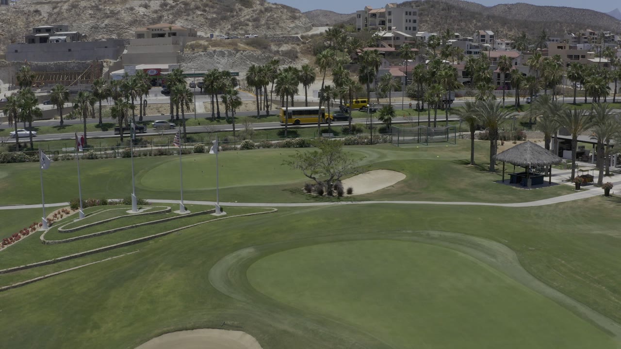 Opening aerial frame over Los Cabos, Mexico, with a large golf green in front, palm-lined roads and buildings beyond, and no clearly identifiable people.