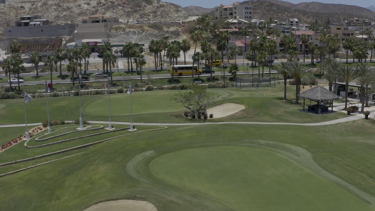 Los Cabos aerial frame with the putting green lower center, a sand bunker to the right, traffic behind the course, and no clearly identifiable people.