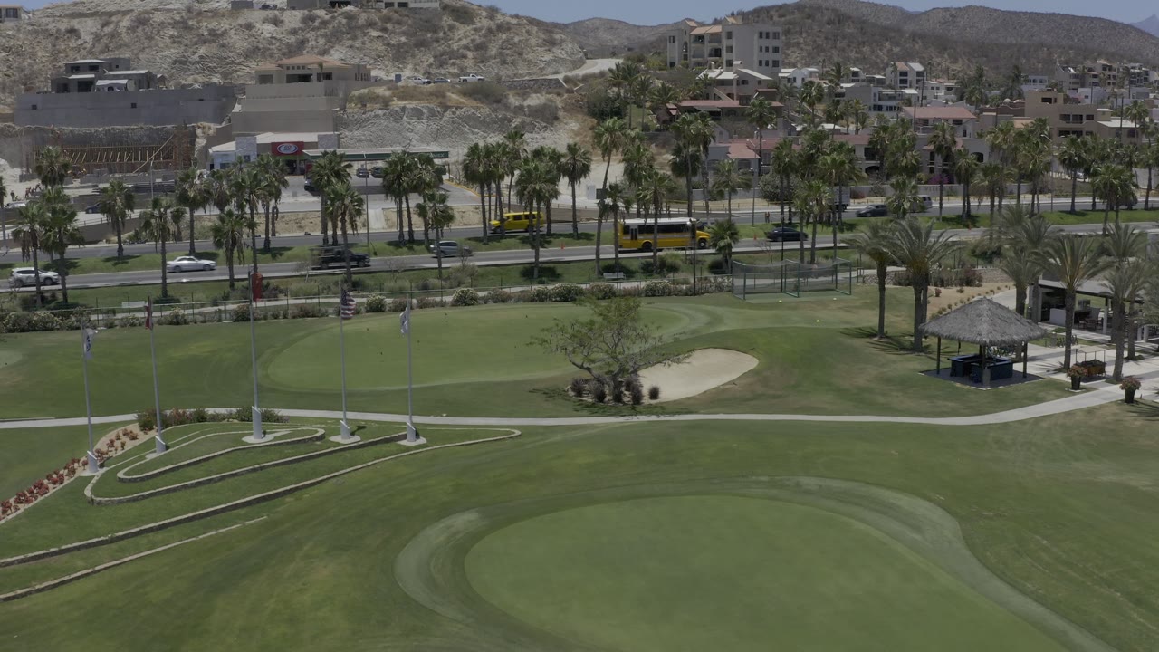 Golf course aerial with flagpoles, curved paths, palms, roads, and hillside buildings.