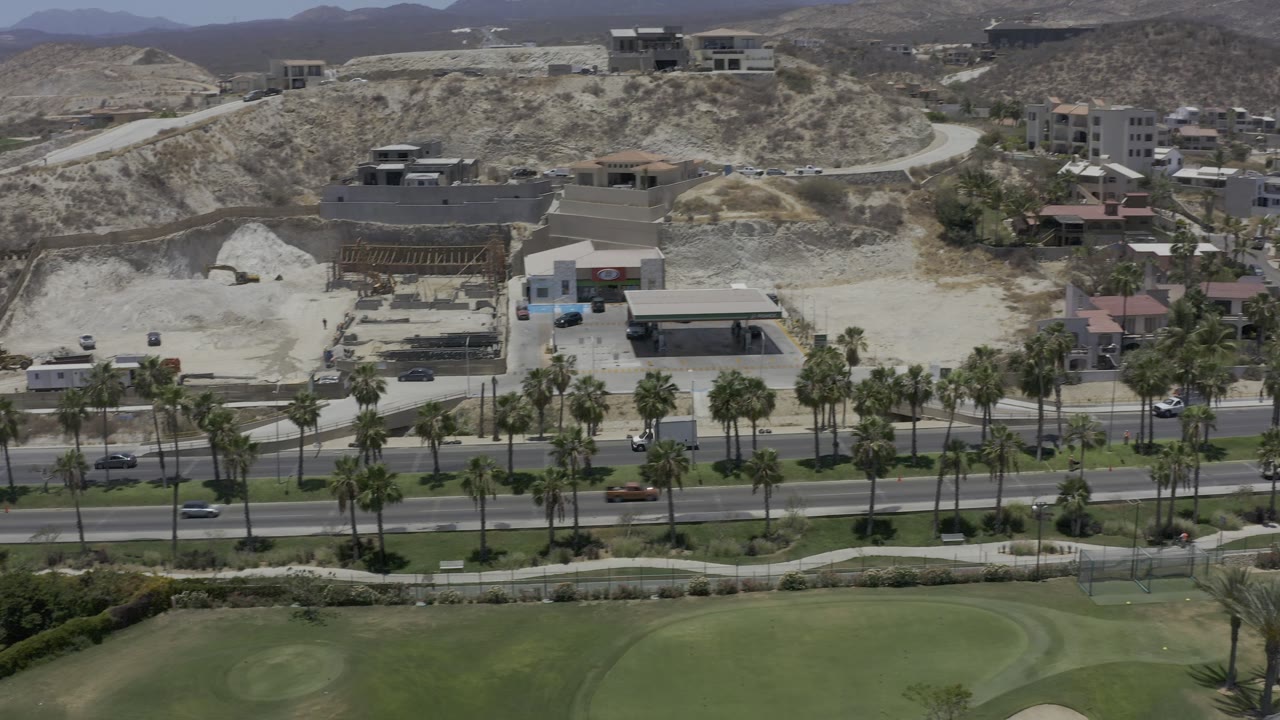 Second Los Cabos aerial frame showing cars along the palm-lined road, a large green lawn at the bottom, hillside buildings, bare earth, and no visible people.