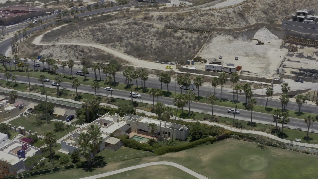 Los Cabos aerial frame with the roadway centered between palm trees, villa roofs and lawns below, and a pale dirt track across the hillside above, with no visible people.