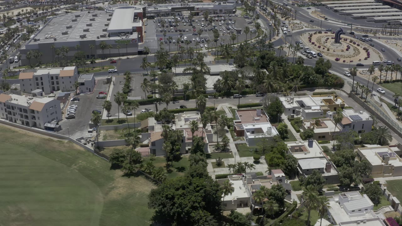 Aerial frame in Los Cabos, Mexico, looking over a residential area toward a curved road, traffic circle, parking lot, and no visible people.