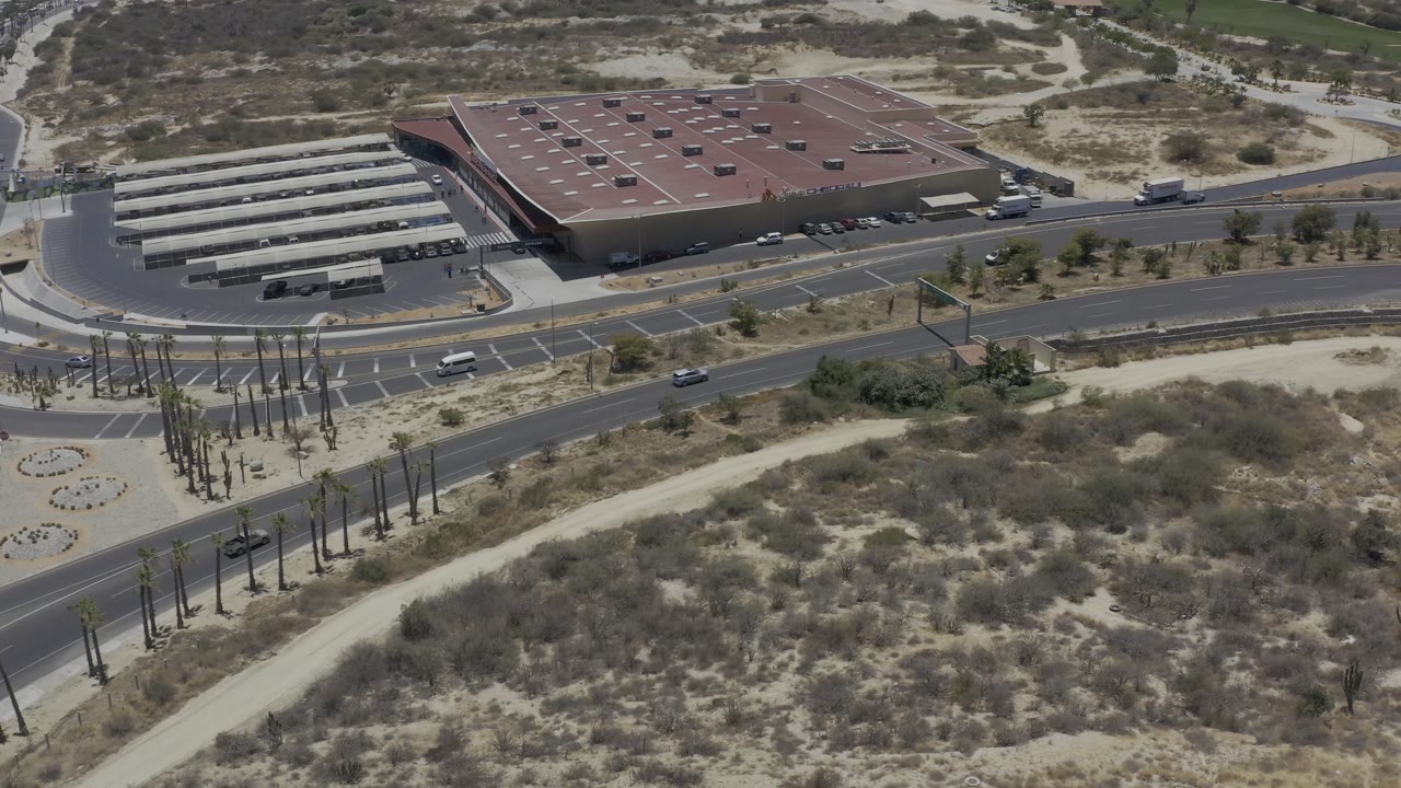 Opening aerial frame over Los Cabos, Mexico, showing a red-roofed commercial building, covered parking rows, intersecting roads, dry brush, and no visible people.