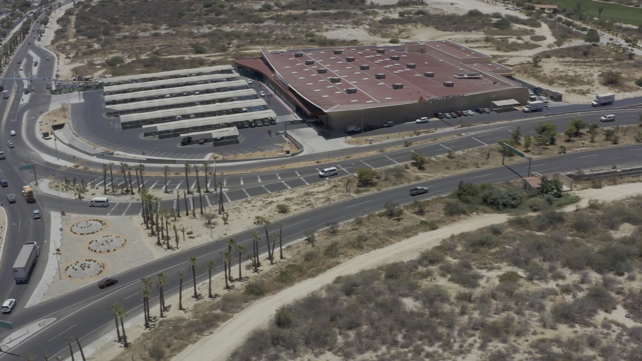 High aerial view over a Los Cabos road junction with a large red-roofed building, rows of parked buses or vehicles, dry scrubland, and no visible people.