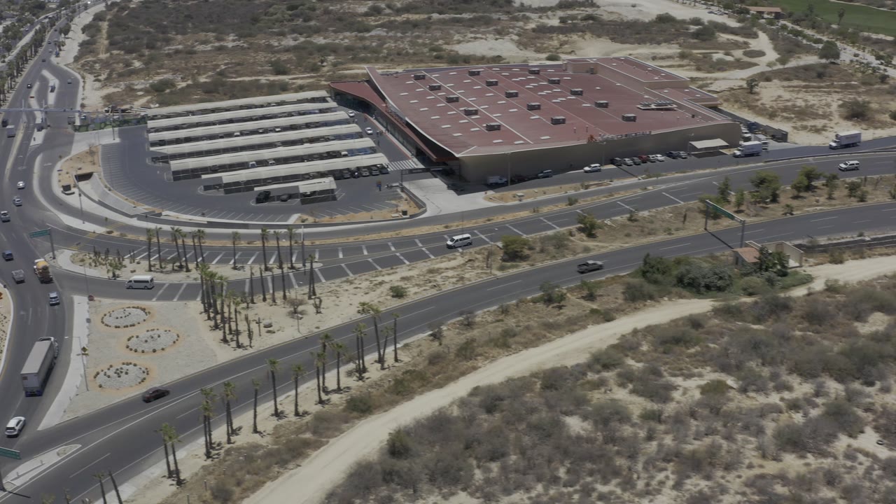 Aerial frame in Los Cabos showing curved roadway lanes, palm-lined medians, a parking lot near a large red-roofed building, dry open land, and no visible people.