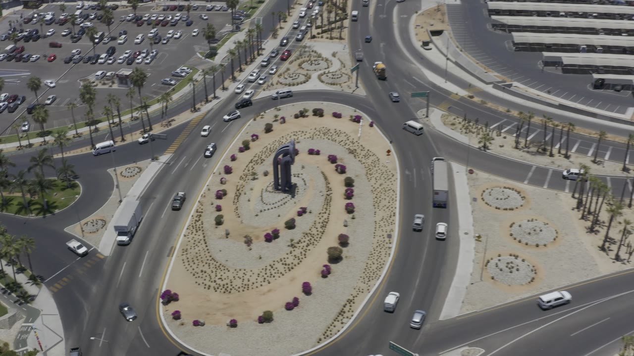 Aerial Los Cabos traffic view with more of the upper parking lot visible, vehicles on several curved lanes, planted medians, and no visible people.
