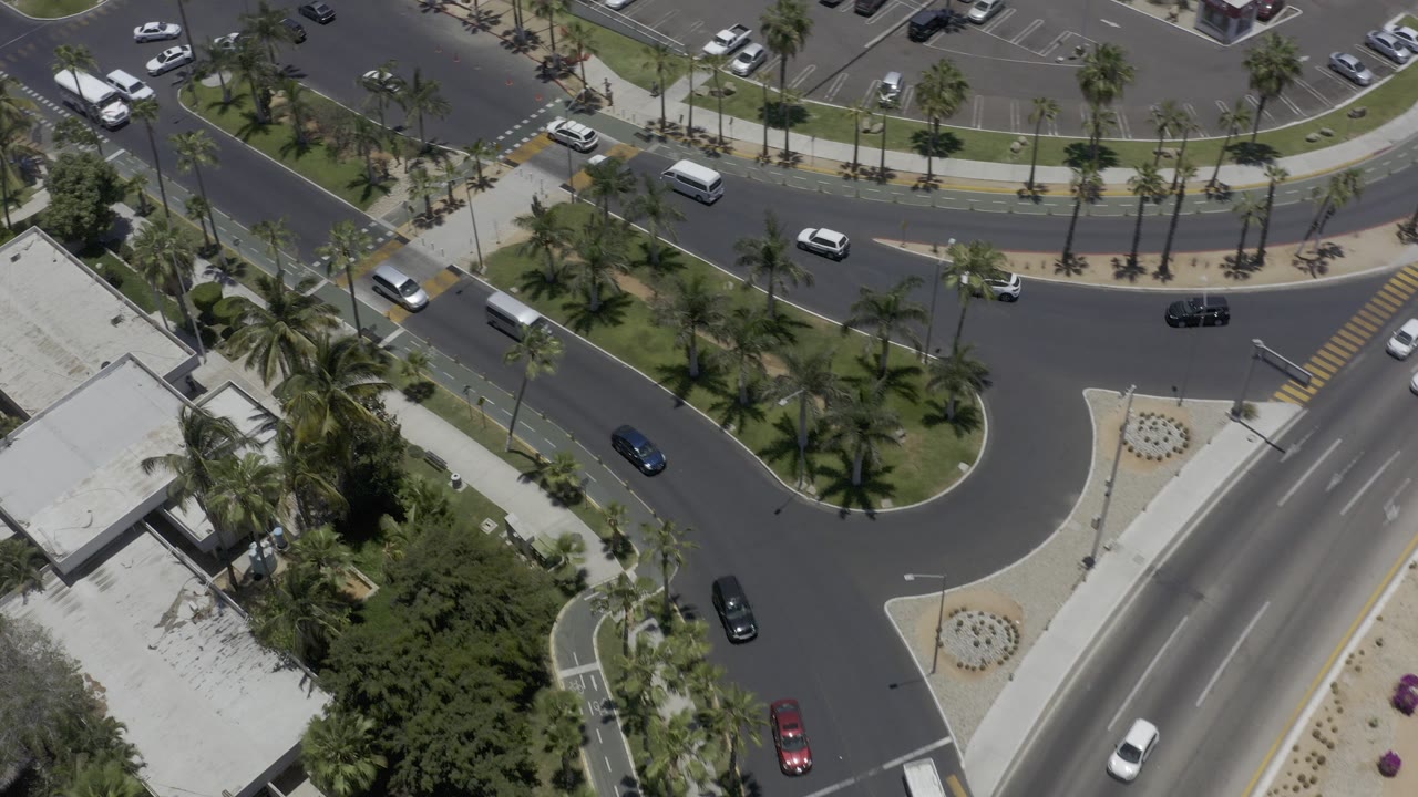 A high overhead view in Los Cabos, Mexico shows curved streets, palm medians, parked cars, and moving vehicles near low buildings, with no visible people.