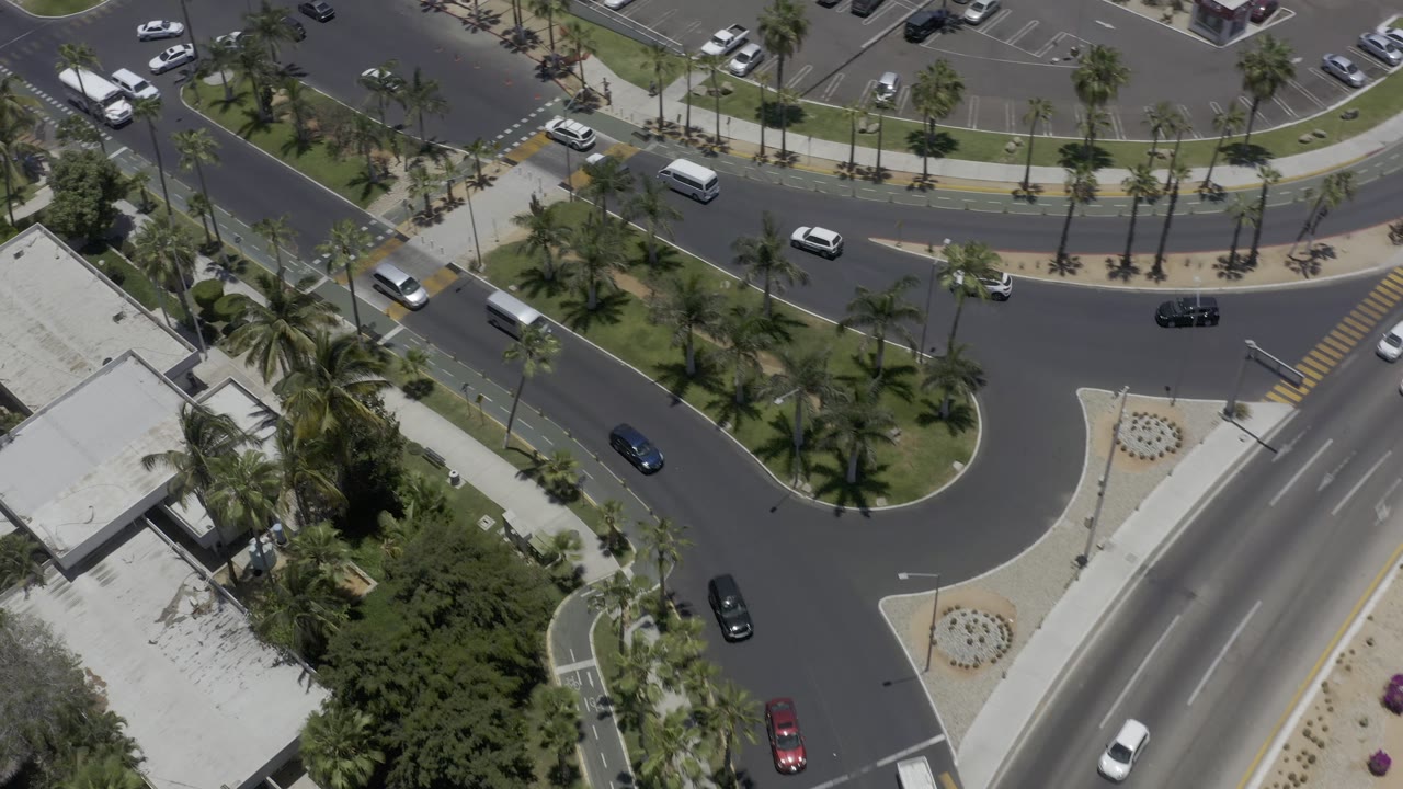 Los Cabos traffic passes through an overhead frame of dark asphalt roads, palm plantings, a crosswalk, parking bays, and white-roofed buildings, with no visible people.