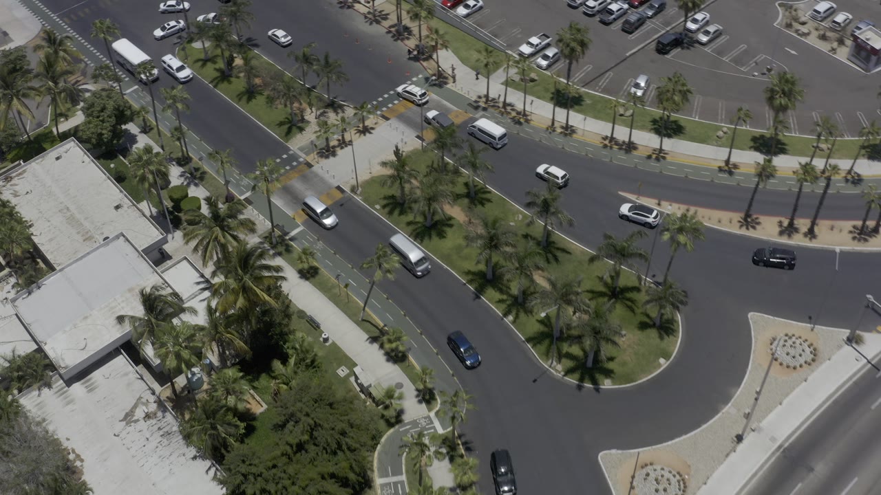 Aerial view of a curved street, parking area, palms, and scattered vehicles.