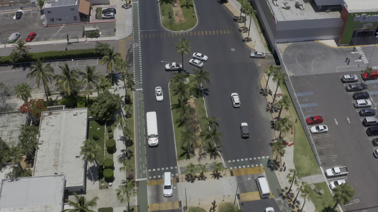 Aerial frame in Los Cabos, Mexico with a white bus or van on the left road, several cars in the intersection, palm medians, parking spaces, and no clearly visible people.