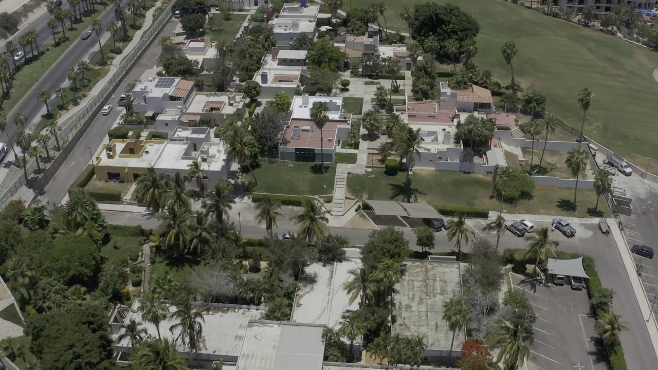 Aerial frame over Los Cabos, Mexico, with flat-roofed homes, palm clusters, a road along the left edge, lawns, and cars near a gated-looking corner, with no visible people.