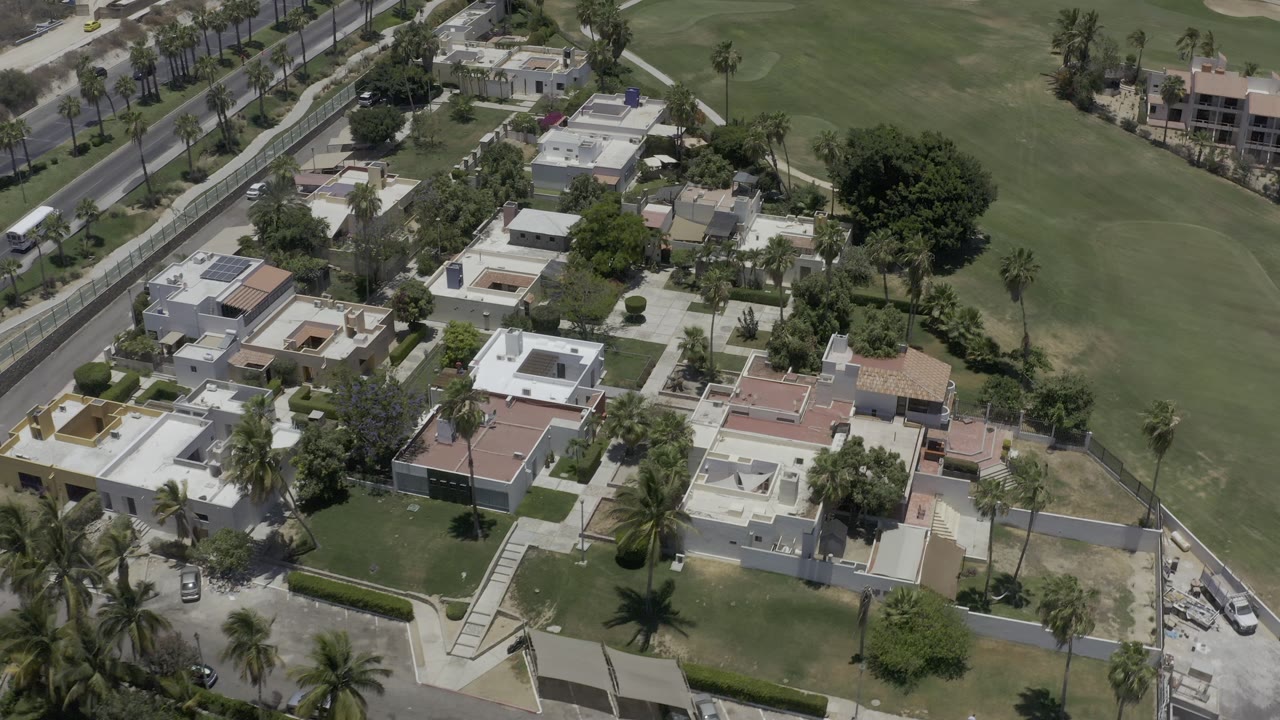 Aerial frame in Los Cabos with white-roofed houses, walled yards, palms, road lanes, and golf-course grass in the background, with no visible people.