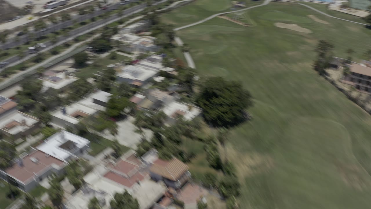 Oblique aerial frame over a Los Cabos golf course with green fairways, low-rise homes, palm-lined streets, and no visible people.