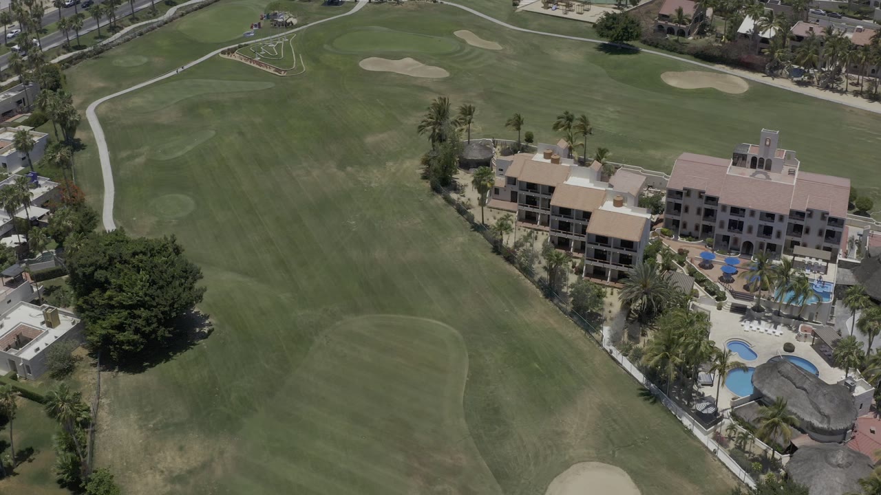 High aerial view in Los Cabos, Mexico, showing green golf fairways, sand bunkers, residential buildings, palm trees, pools, and no visible people.