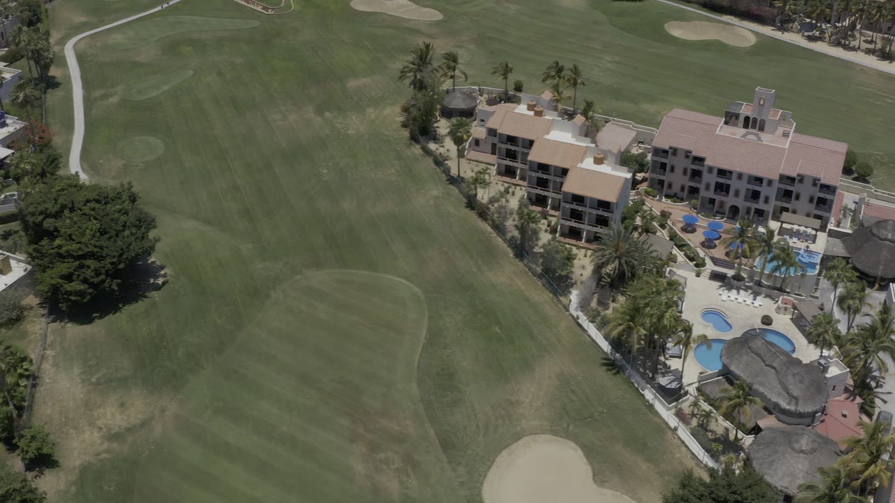 Aerial view in Los Cabos, Mexico showing a broad green fairway, sand bunkers, palm trees, resort-style buildings, blue pools, and no visible people.