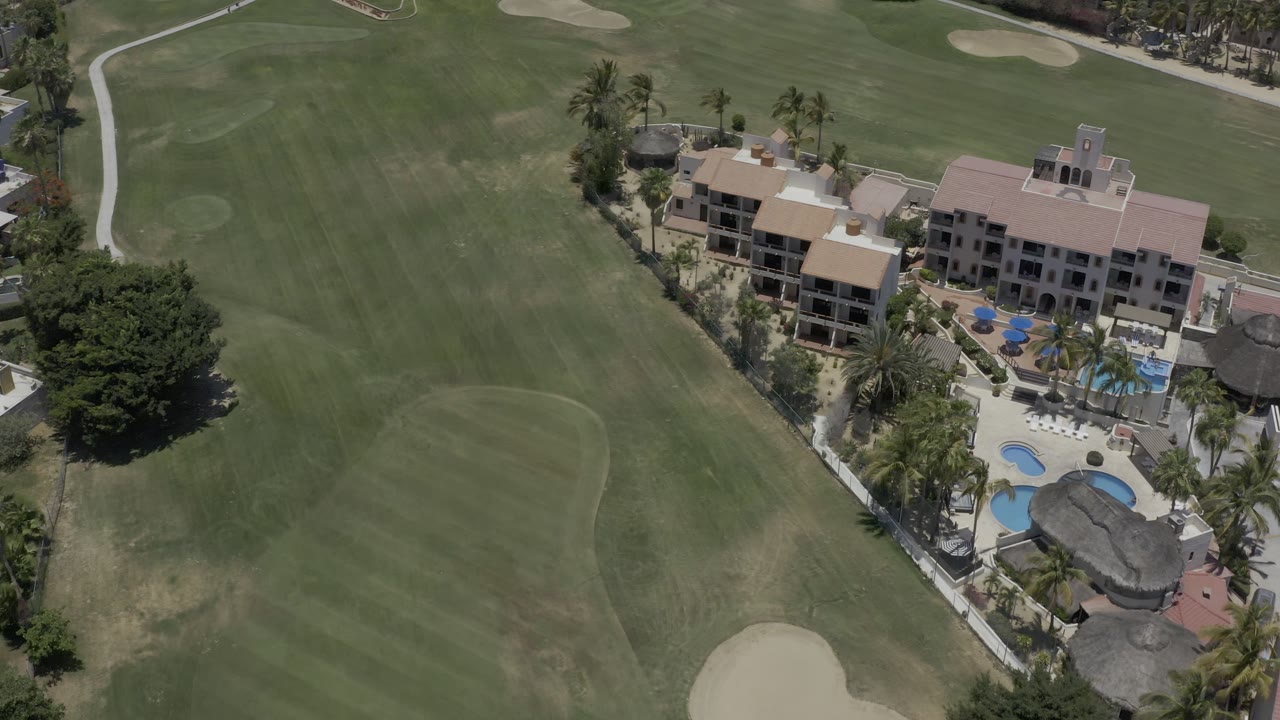 A second aerial frame in Los Cabos, Mexico keeps the resort buildings centered beside fairway grass, pale bunkers, pool shapes, palms, and no visible people.