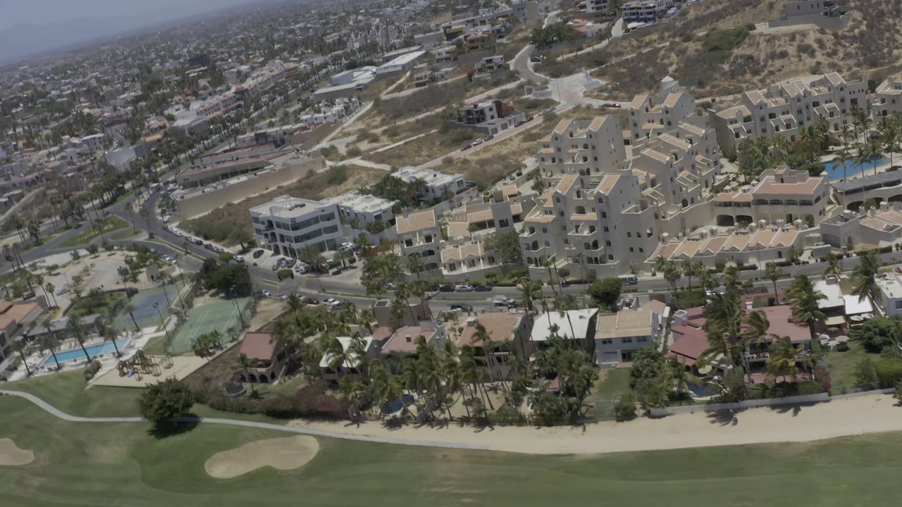 Wide aerial frame in Los Cabos, Mexico, with beige apartment blocks, curved roads, villas, blue pools, golf-course grass, palms, hazy distance, and no visible people.