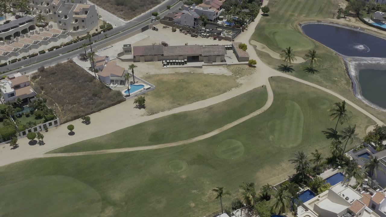 Aerial frame over Los Cabos showing the golf green and cart path lower in frame, palms near the homes, a road at the top, and the water feature along the right edge, with no visible people.