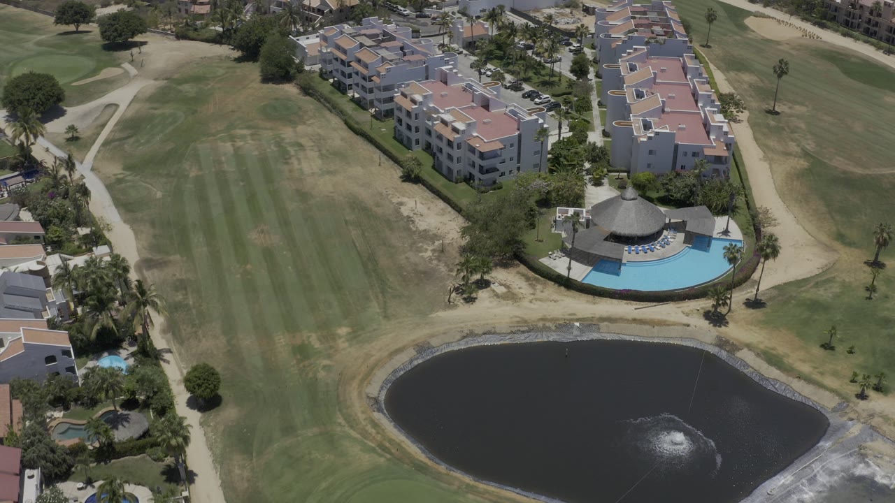 Aerial frame over a Los Cabos residential complex with terracotta-roof buildings, a curved swimming pool, open grass, paths, and a dark pond in the foreground, with no visible people.