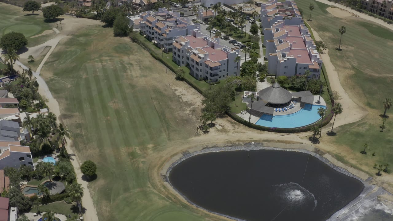 Aerial view in Los Cabos showing the pool pavilion area, low-rise residences, dry and green grass bands, curving paths, parked cars, and a pond fountain, with no visible people.