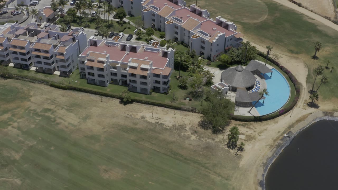 Aerial view of resort buildings, golf course, curved pool, paths, and dark water edge.
