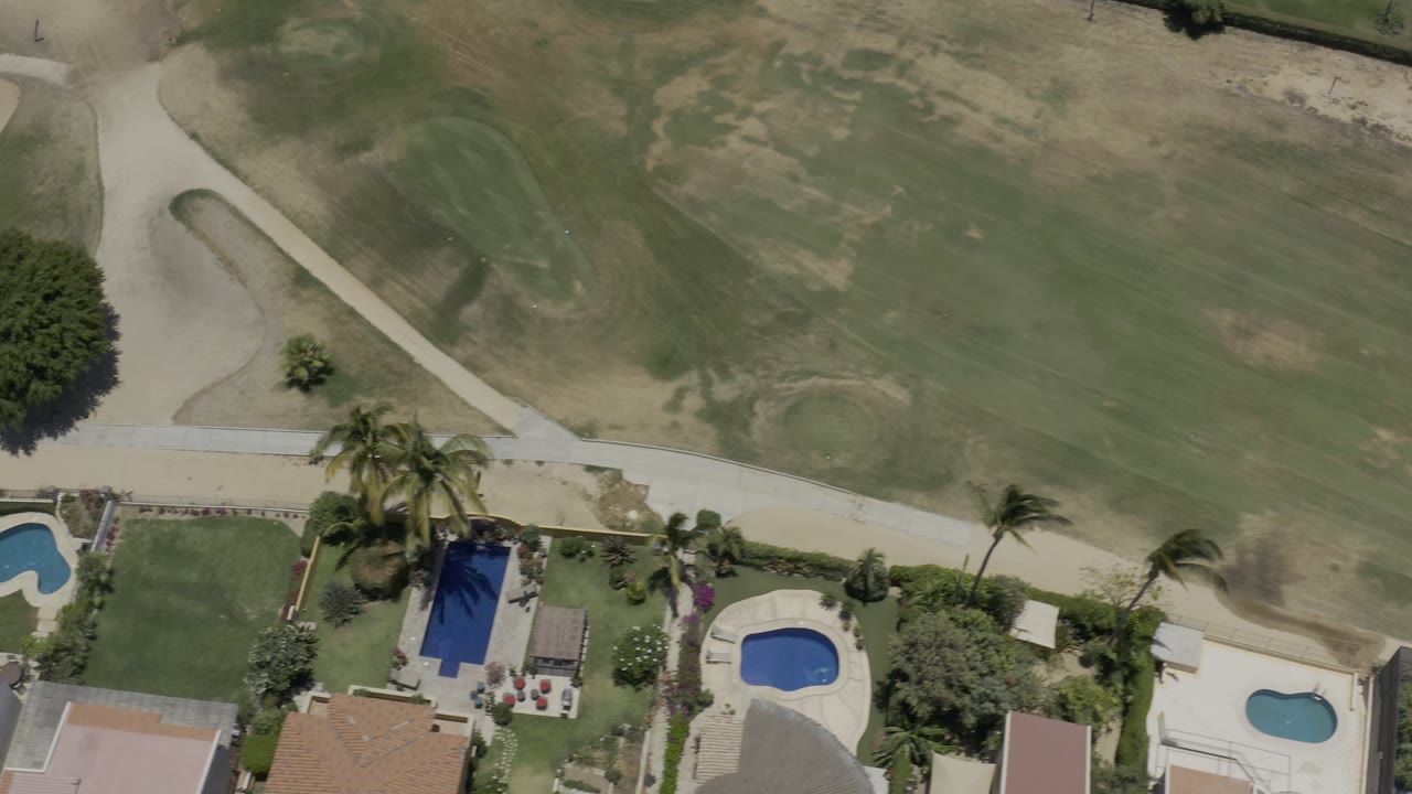 Aerial Los Cabos view with two blue pools near tiled roofs, palms, a pale path, and golf course grass filling the upper half, with no visible people.