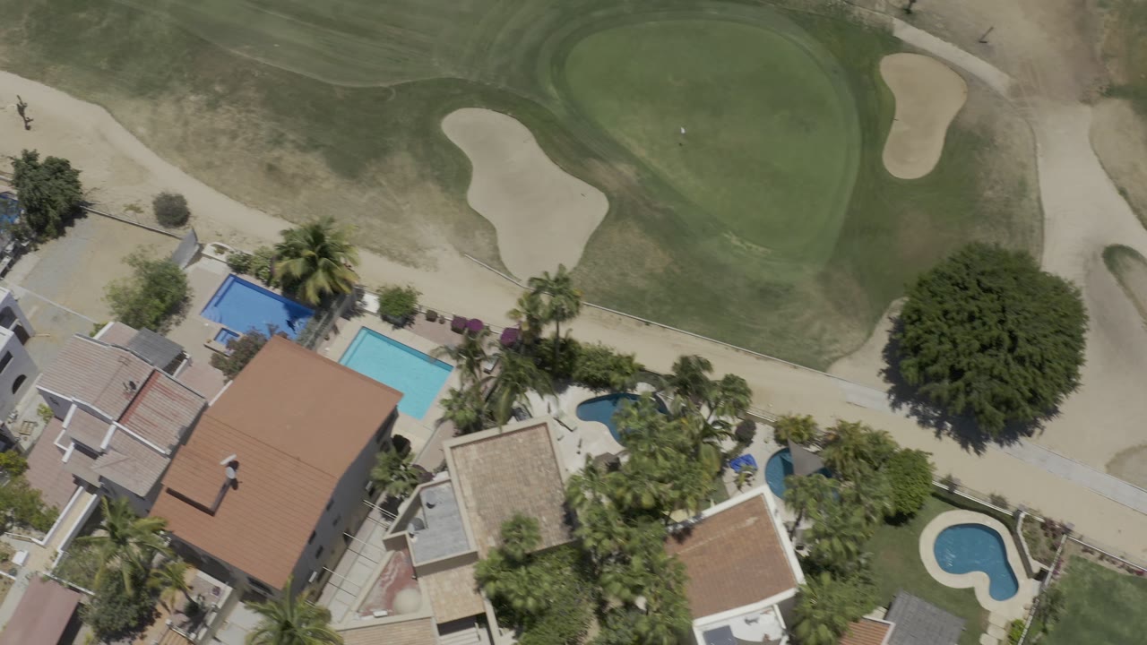 Los Cabos overhead view with a large red-roofed house, bright rectangular pool, curved backyard pool, palm trees, golf turf, and pale sand bunkers, with no visible people.