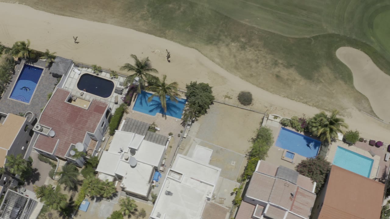 Top-down aerial frame from Los Cabos, Mexico showing several homes with blue pools beside a sandy path and green lawn, with no clearly identifiable people visible.