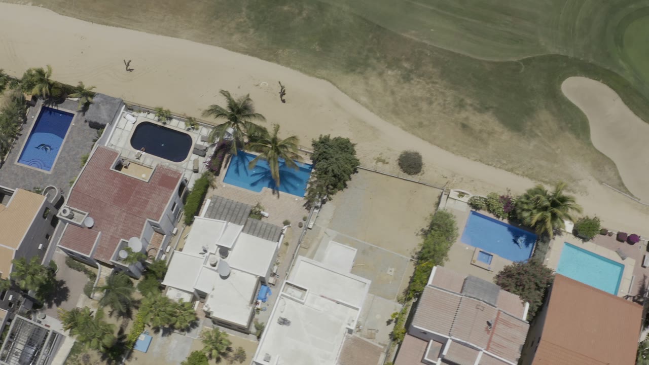 High aerial frame in Los Cabos, Mexico with red and white rooftops, two dark and blue pools, palms, sandy walkway, and no clearly identifiable people visible.