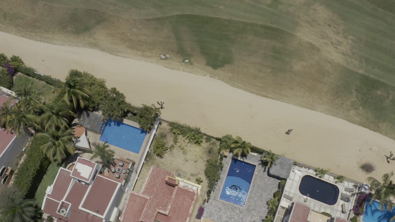Top-down aerial view of homes, pools, palms, sandy path, and grass.