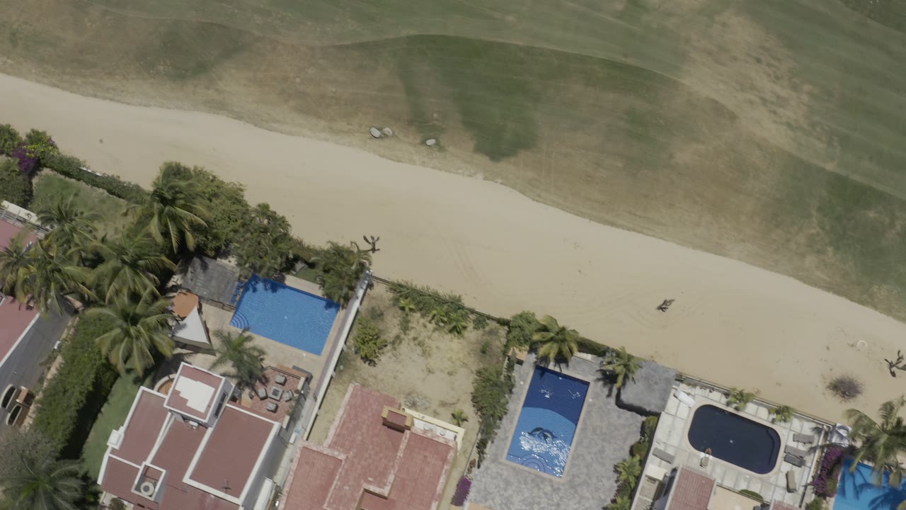 Overhead Los Cabos view with the sandy strip running across the frame, grass above it, residential roofs and blue pools below it, and no visible people.