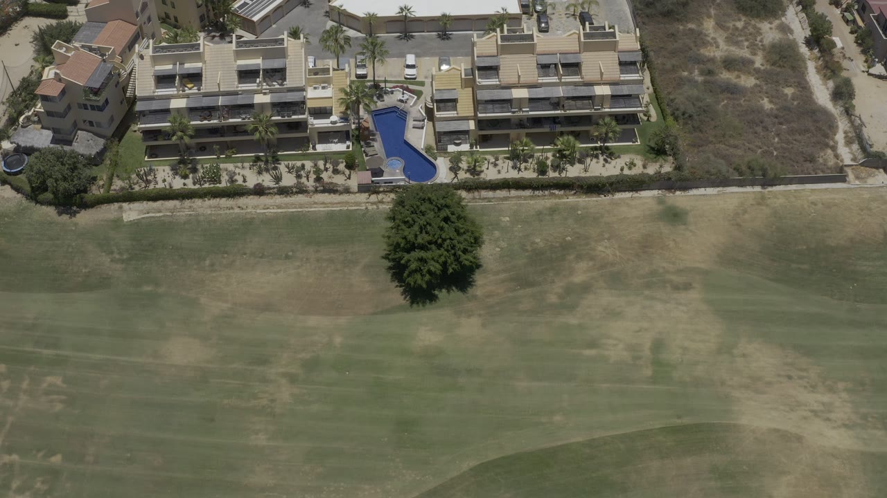 Los Cabos aerial view with the same resort complex near the top edge, a blue pool at center right, scrubby open ground, and golf turf filling the lower frame with no visible people.