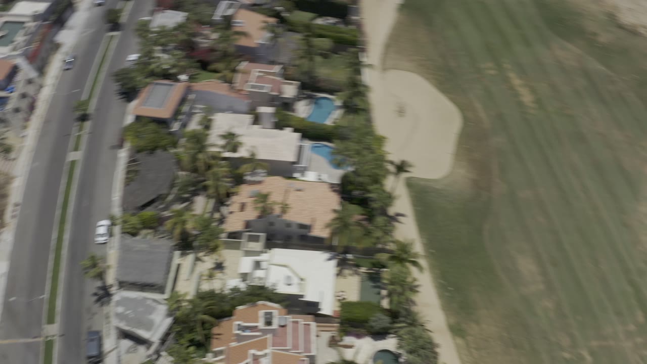 A second overhead frame in Los Cabos, Mexico, centers walled homes, bright blue pools, palm trees, a road, and green landscaped ground, with no visible people.