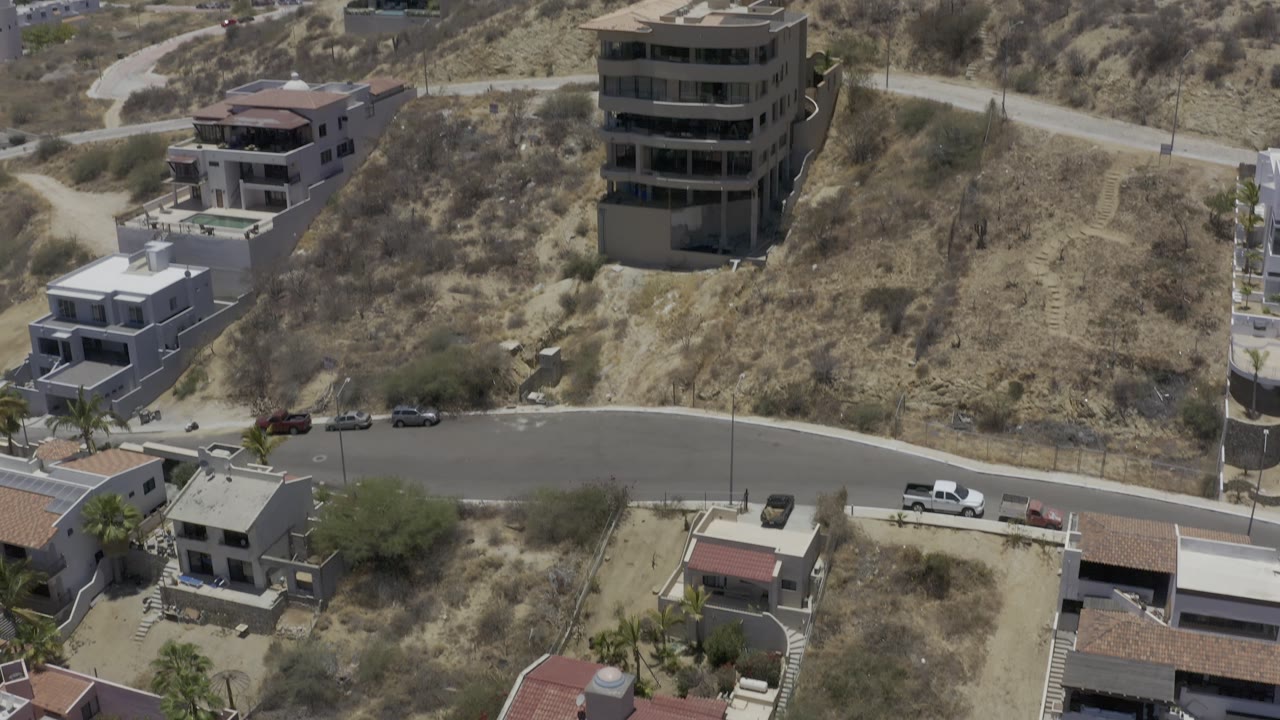 Aerial frame in Los Cabos, Mexico, with a paved road crossing below modern hillside homes, parked cars, dry slopes, and no visible people.